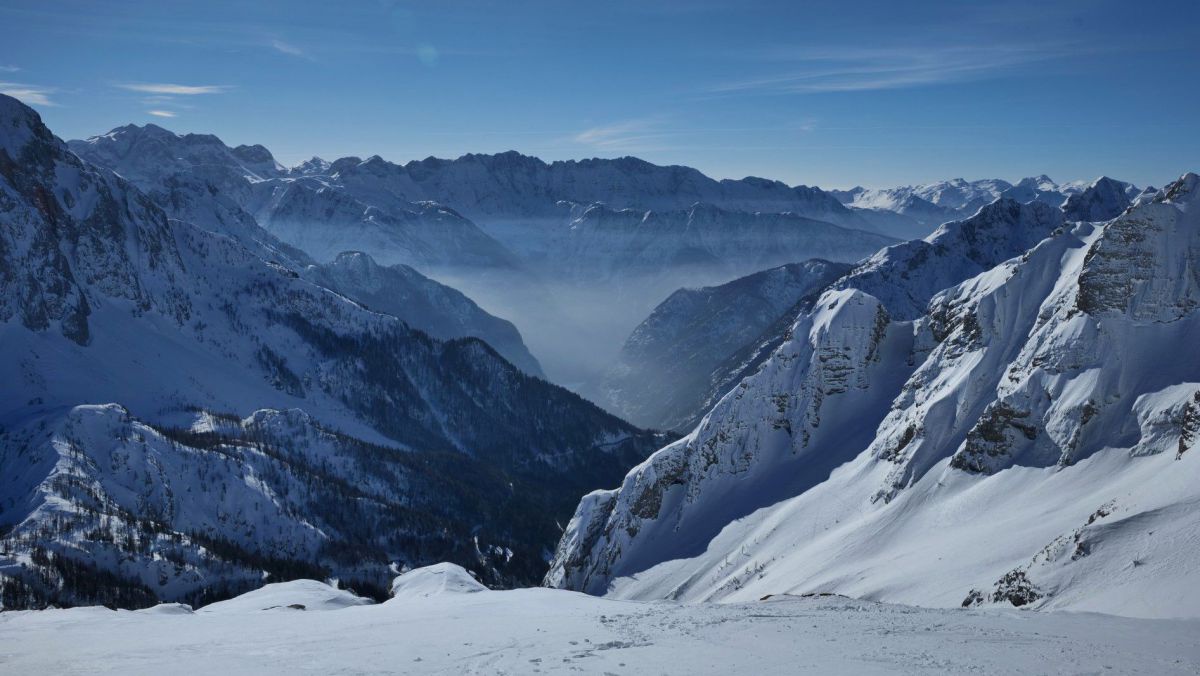 Bergpanorama soweit das Auge reicht, Nebel hängt zwischen niederen Gipfeln, darüber strahlend blauer Himmel und sonnenbeschienene Schneeflächen.